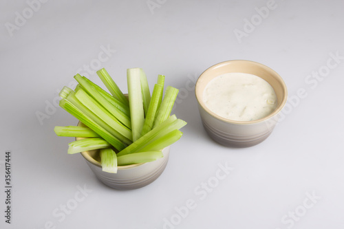 close up isolated top corner view shot of a bowl of crunchy juicy green celery sticks next to a white cup of blue cheese dipping sauce on a white background
