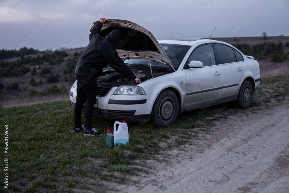 Man standing near car with open hood on sunset. Car breakdown. Confused man stands with open hood with selective soft focus. Man standing near car with open hood on sunset. Car breakdown. Confused man stands with open hood with selective soft focus.