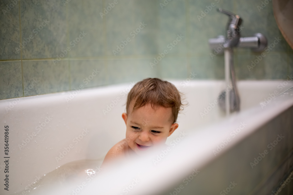 Cute Caucasian baby take a bath, playi in bath and smiles looking at water splashes, in the background a green bathroom in blur. close-up, soft focus Cute Caucasian baby take a bath, playi in bath and smiles looking at water splashes, in the background a green bathroom in blur. close-up, soft focus