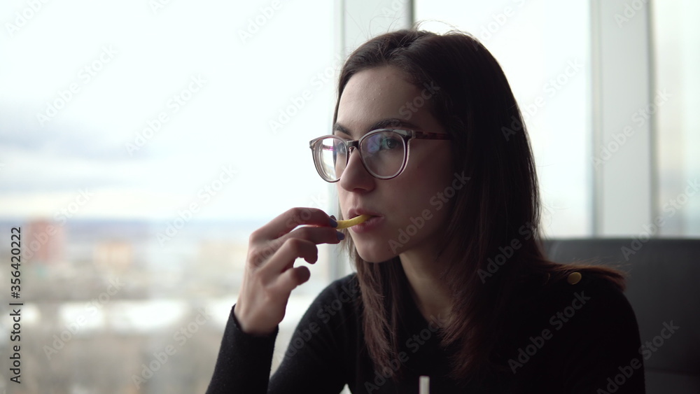 Obraz premium Young woman eating fried potatoes. A girl sits in a cafe by the panoramic window and eats fast food.