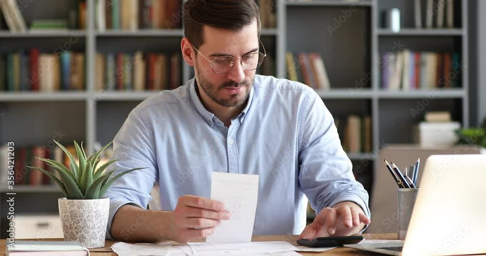 Focused young businessman in glasses holding paper receipt, doing accounting at office. Concentrated bearded man calculating household bills, managing monthly incomes and outcomes alone at home. Focused young businessman in glasses holding paper receipt, doing accounting at office. Concentrated bearded man calculating household bills, managing monthly incomes and outcomes alone at home.