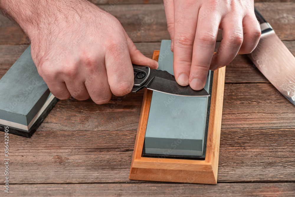 Man sharpens his knives with a whetstone on a rustic wooden table Man sharpens his knives with a whetstone on a rustic wooden table