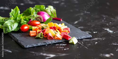 Uncooked colorful pasta in shape of heart and fresh vegetables and greens on black slate board. Copy space photo