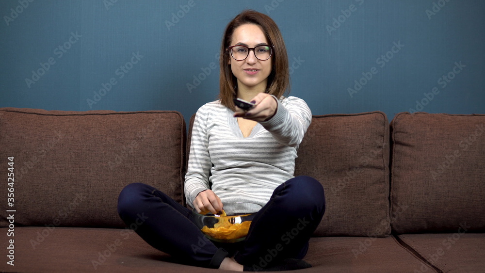 A young woman in glasses eats chips and watches TV. Girl sitting with legs on the couch A young woman in glasses eats chips and watches TV. Girl sitting with legs on the couch