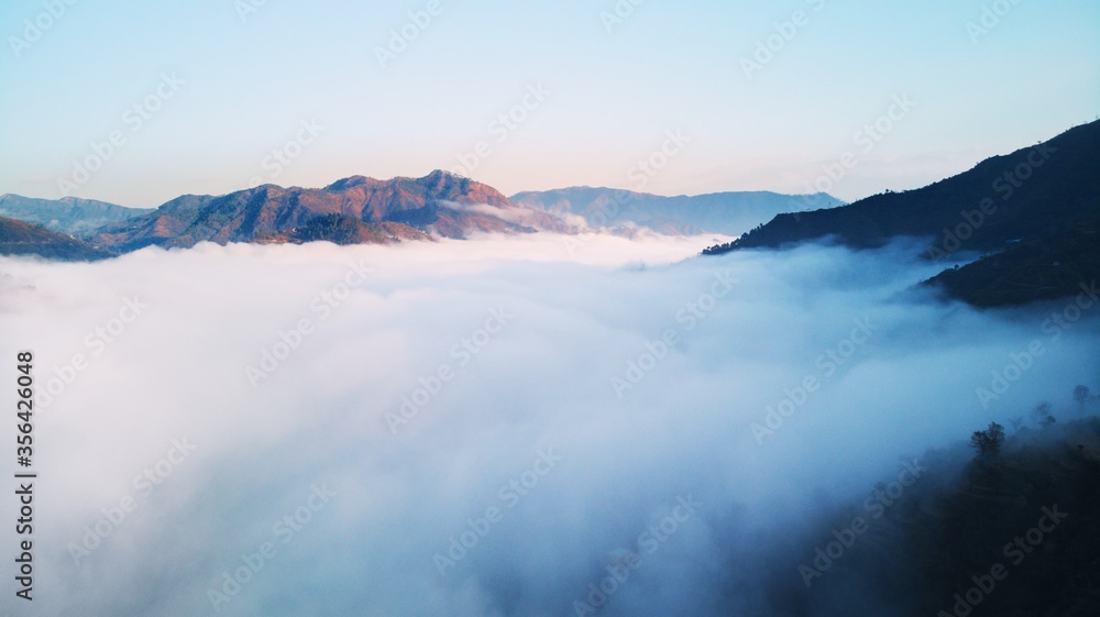 Aerial/Drone View of a foggy Himalayan valley and green fields surrounded by lush green hills during a beautiful morning at a rural location in Uttarakhand, India.