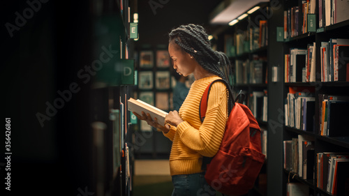 University Library: Portrait of Gifted Beautiful Black Girl Stands Between Rows of Bookshelves and Searching for the Right Book Title, Finds and Picks one for Class Assignment photo