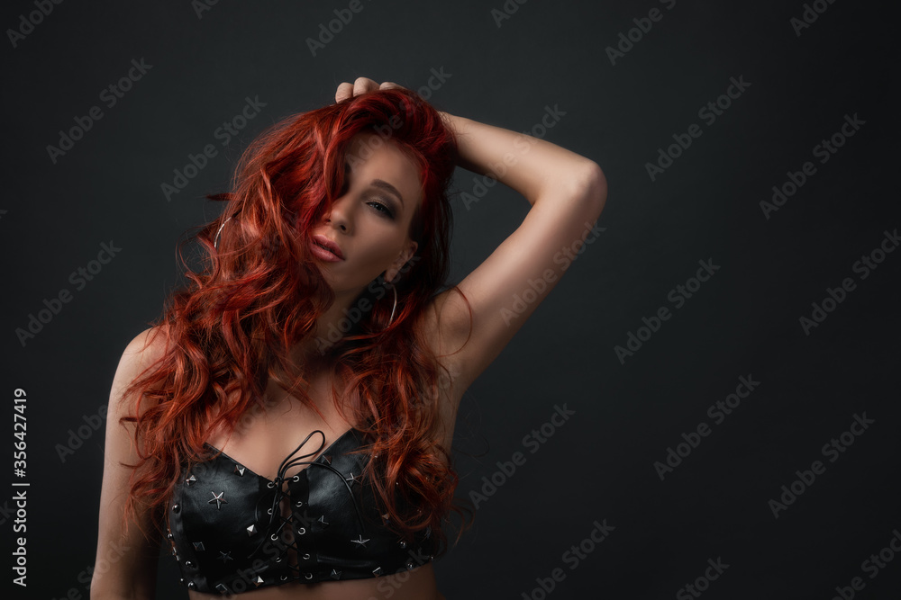 Fototapeta premium concept of hair coloration and skin care with cosmetics, copy space. sexy girl touching her red colored hair. woman with professional make up wearing leather top bra with sparkling stars.