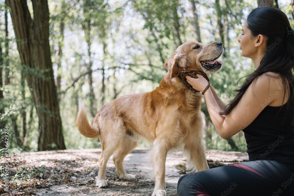 Pretty lady petting her dog in forest. Side view of girl in sport outfit sitting in nature and caressing her dog during summer time.