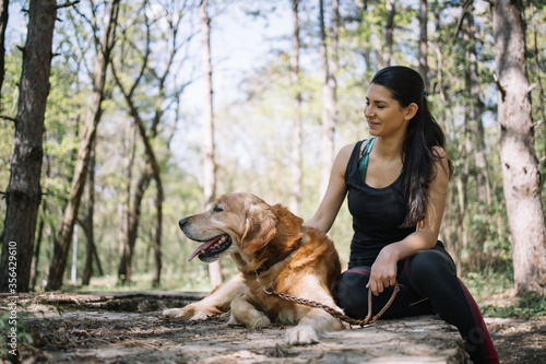 Woman and her dog sitting in park. Brunette girl dressed with sporting clothes sitting next to her dog in nature.