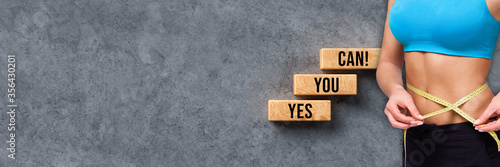 woman measuring her body in front of wooden blocks with message YES YOU CAN on concrete background