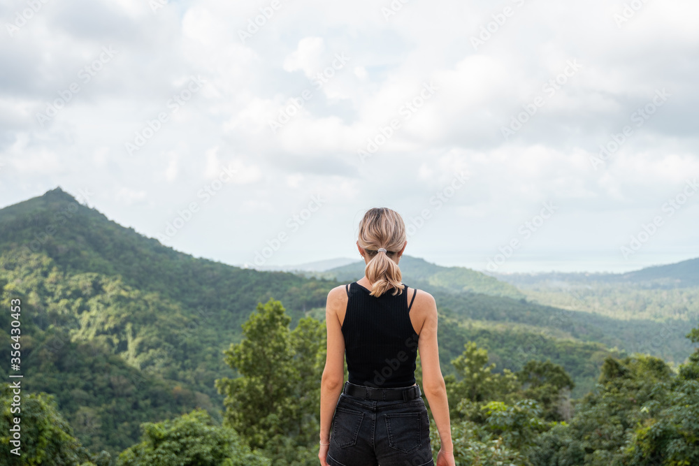 Back side of woman in black t-shirt enjoying tropical forest view with mangrove trees. Mountains and white clouds on a blue sky. Tropical summer holiday vacation concept. Back side of woman in black t-shirt enjoying tropical forest view with mangrove trees. Mountains and white clouds on a blue sky. Tropical summer holiday vacation concept.