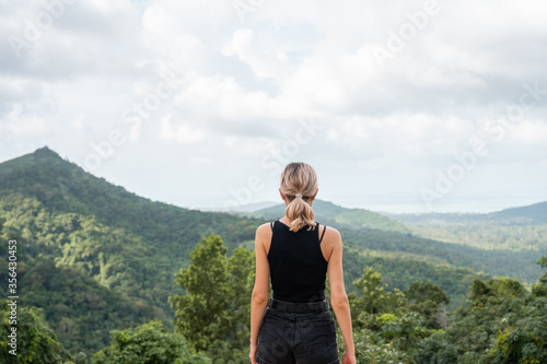 Back side of woman in black t-shirt enjoying tropical forest view with mangrove trees. Mountains and white clouds on a blue sky. Tropical summer holiday vacation concept.