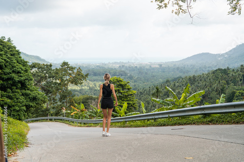 Woman in black t-shirt enjoying tropical forest view while standing on a empty road. Mountains and white clouds on a blue sky. Tropical summer holiday vacation concept.