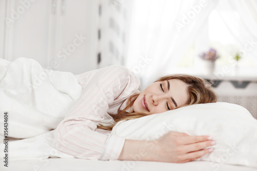 An attractive young girl in striped pajamas is lying with her eyes closed on the bed in her bright bedroom in the morning. Side view