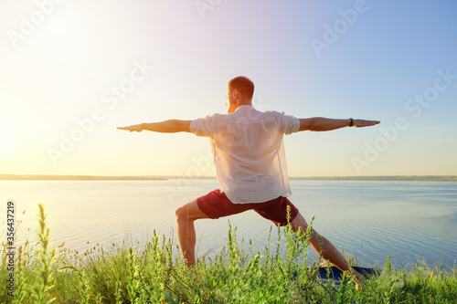 Fit muscular man doing yoga by the water on the grass with hands stretch. Orange sunlight sunset tranquility at lake. Male back in white office shirt. Pilates warrior pose Healthy spine workout