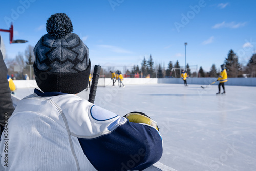 Young Hockey Player watching outdoor ice hockey game from behind the boards