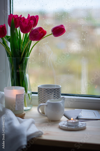 Still life coffee at home, a cozy coffee, tulips in a vase. Coffee on the windowsill, comfort at home. Milk jug  mug, candlestick, envelope and postcard on wooden desk. Spring content