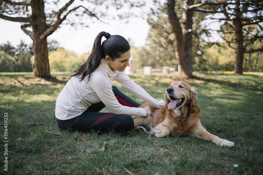 Woman in sportswear petting her retriever dog. Pretty girl and dog lying on ground in park near trees during sunny day. Woman in sportswear petting her retriever dog. Pretty girl and dog lying on ground in park near trees during sunny day.