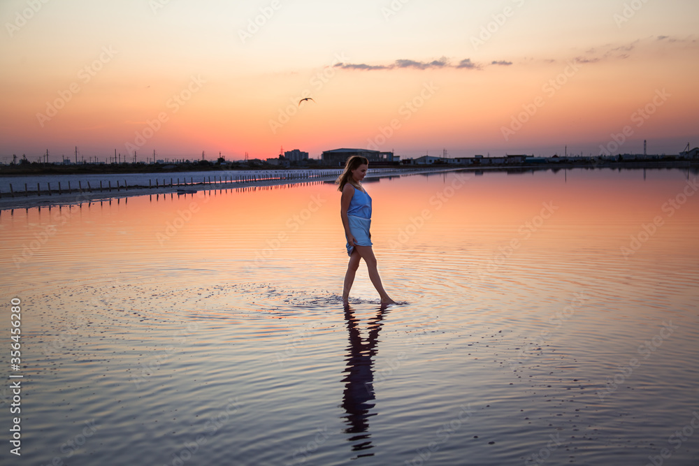 Fototapeta premium Rear view of the woman back silhouette look to the purple sunrise in water on summer. Woman walking on the shore of the salt beach at pink lake. vacation lifestyle near ocean