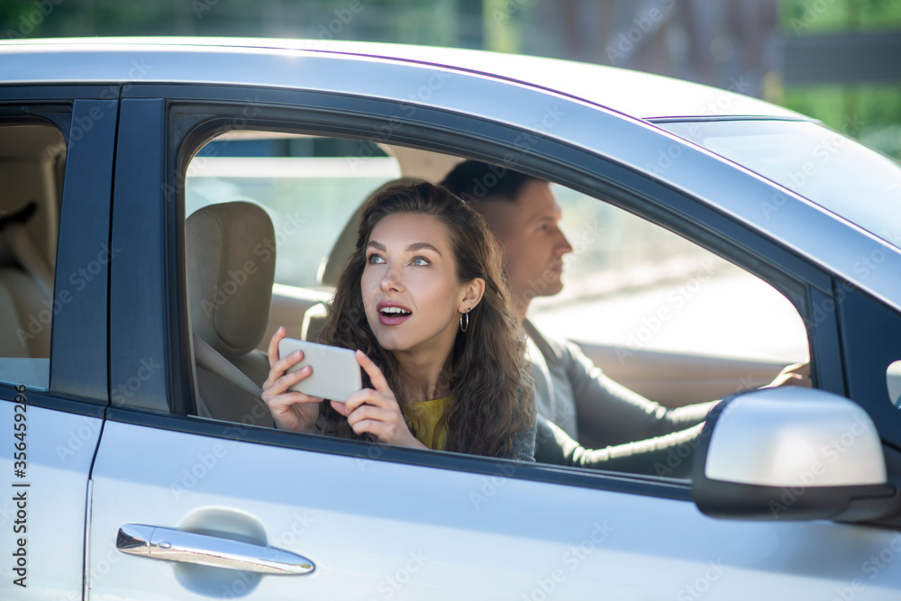 Young couple sitting in the car, woman making photos and looking surprised