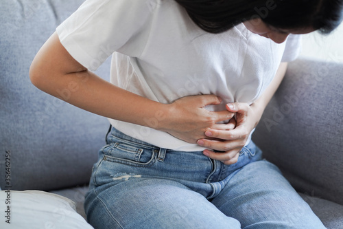 Young Asian woman suffering from strong abdominal pain stomachache or period comes while sitting on sofa at home