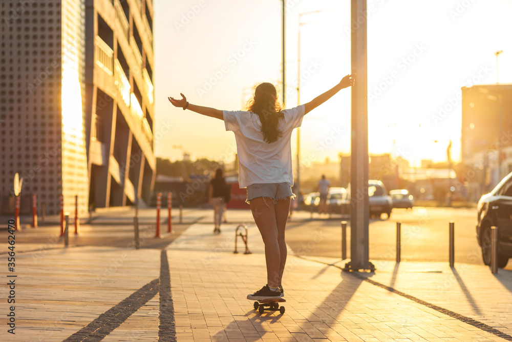 Girl on a skateboard illuminated by the rays of the setting sun. rides on a skateboard with his arms raised high, his back to the camera. Selective focus. Sunset in the city Girl on a skateboard illuminated by the rays of the setting sun. rides on a skateboard with his arms raised high, his back to the camera. Selective focus. Sunset in the city