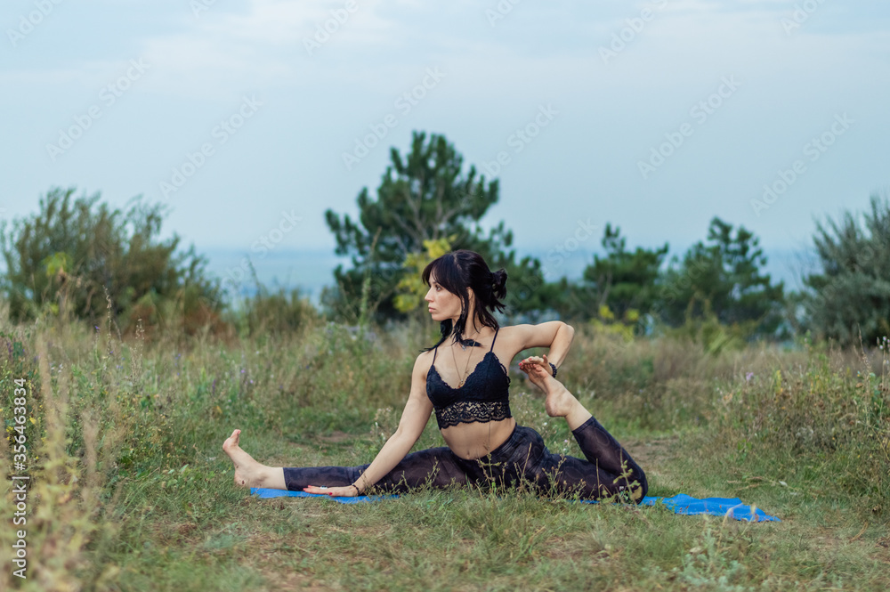 A dark-haired beautiful flexible girl in a black top and leggings practices yoga on a blue Mat in nature. Beautiful scenery. Pines.
