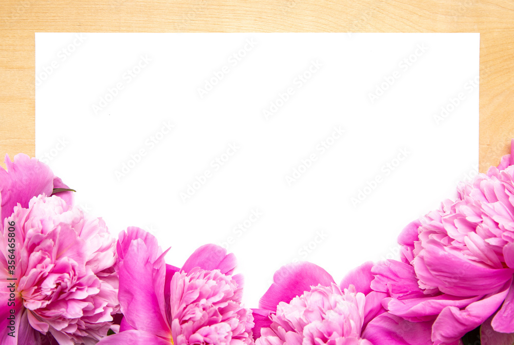 White paper card on rustic wooden background with pink peonies. Flowers. Workspace. Blank for greetings
