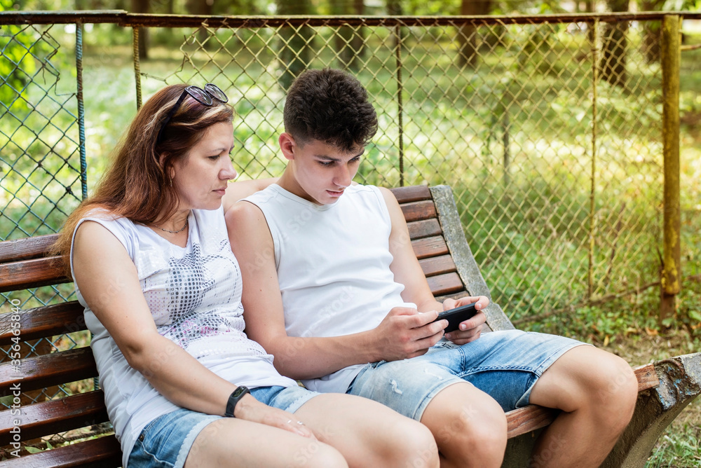 Teenager sits on a park bench and plays games on the phone, mom sits next to him and also looks at the phone, she wants to talk to her son and he is busy Teenager sits on a park bench and plays games on the phone, mom sits next to him and also looks at the phone, she wants to talk to her son and he is busy