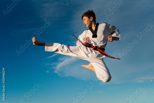 Karate boy kicks in the air and flies over the blue background of the sky. Practicing Taekwondo in nature