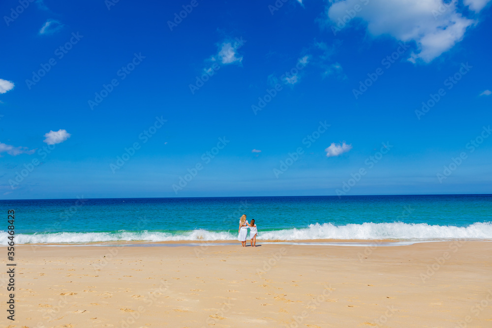 Young beautiful mother with her charming daughter in white clothes walk on a sandy beach near the water
