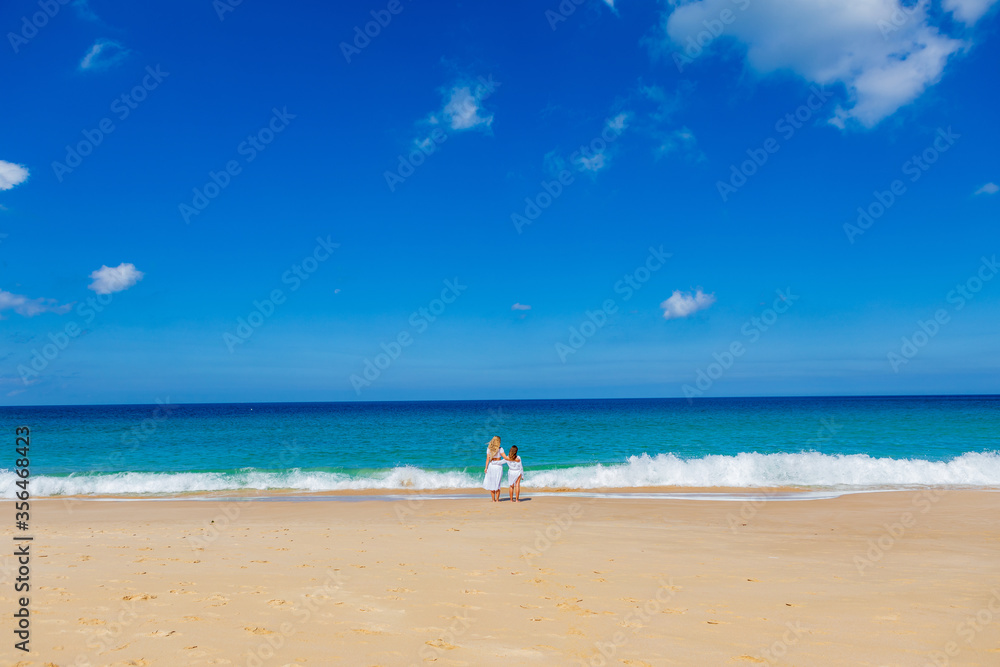 Young beautiful mother with her charming daughter in white clothes walk on a sandy beach near the water Young beautiful mother with her charming daughter in white clothes walk on a sandy beach near the water
