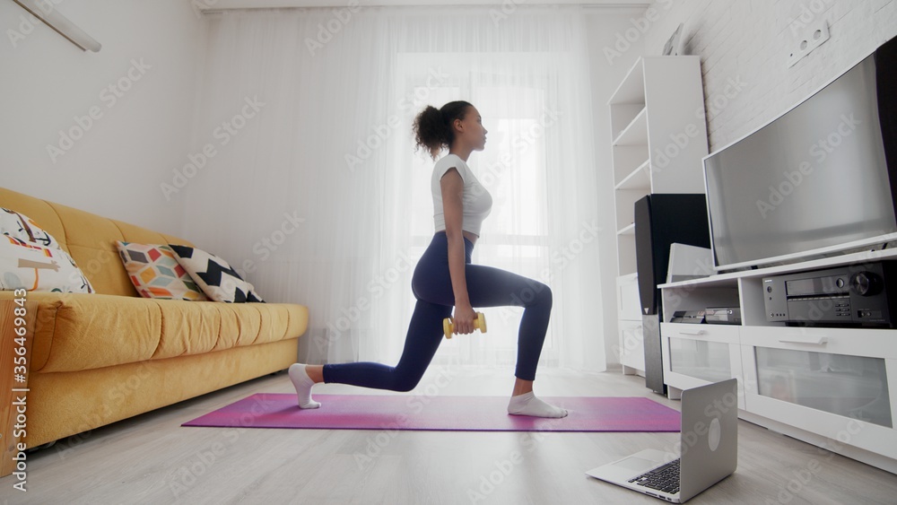 On-line home work out woman using internet services with help of her instructor on laptop at home. Slim sporty african american woman does split squats with dumbbells On-line home work out woman using internet services with help of her instructor on laptop at home. Slim sporty african american woman does split squats with dumbbells