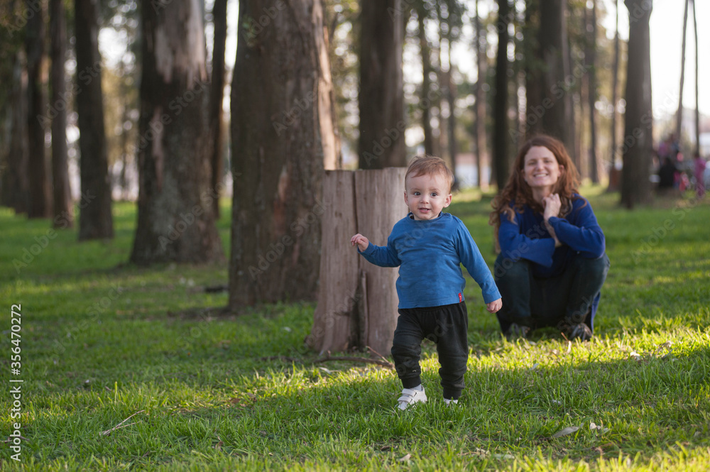 Fototapeta premium A young mother smiles at her one year and a haly years old son as he walks through a park.
