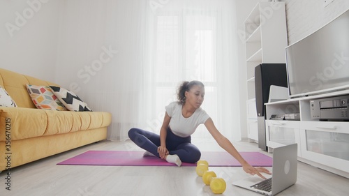 On-line home work out woman using internet services with help of her instructor on laptop at home. Slim sporty african american woman sits in lotus pose and uses the laptop