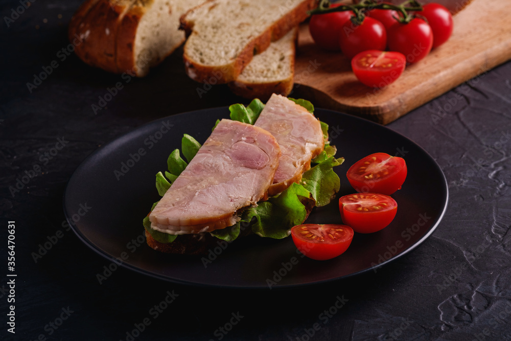 Sandwich with turkey ham meat, green salad and fresh cherry tomatoes slices on black plate near to ingredients on cutting board, dark textured background, angle view