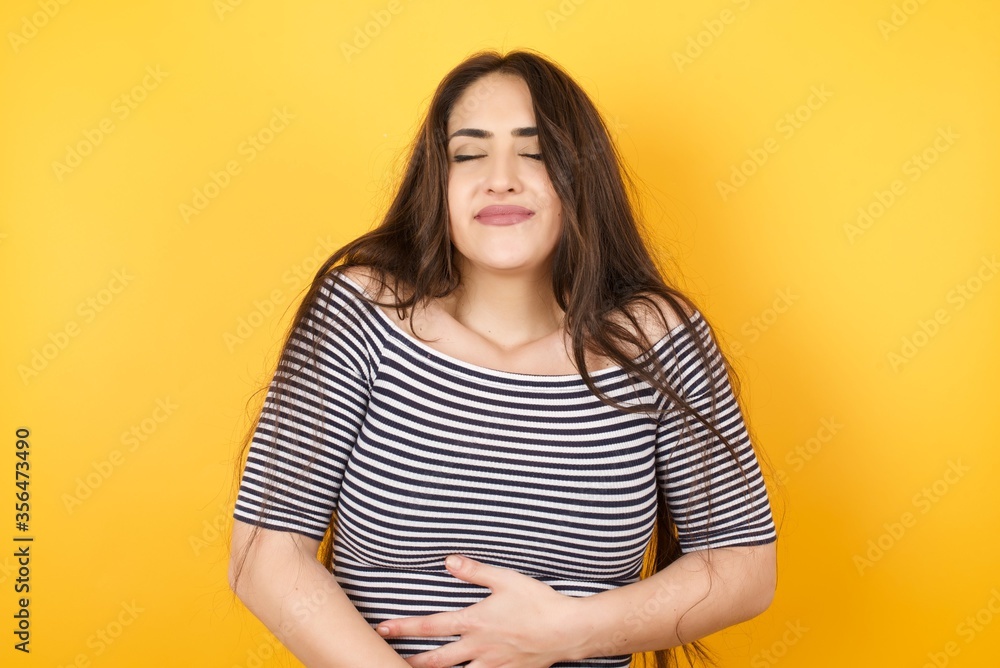 Satisfied smiling young woman keeps both hands on belly, being in good mood after eating delicious supper, demonstrates she is full, isolated over yellow background. Pleasant feeling in stomach
Satisfied smiling young woman keeps both hands on belly, being in good mood after eating delicious supper, demonstrates she is full, isolated over yellow background. Pleasant feeling in stomach