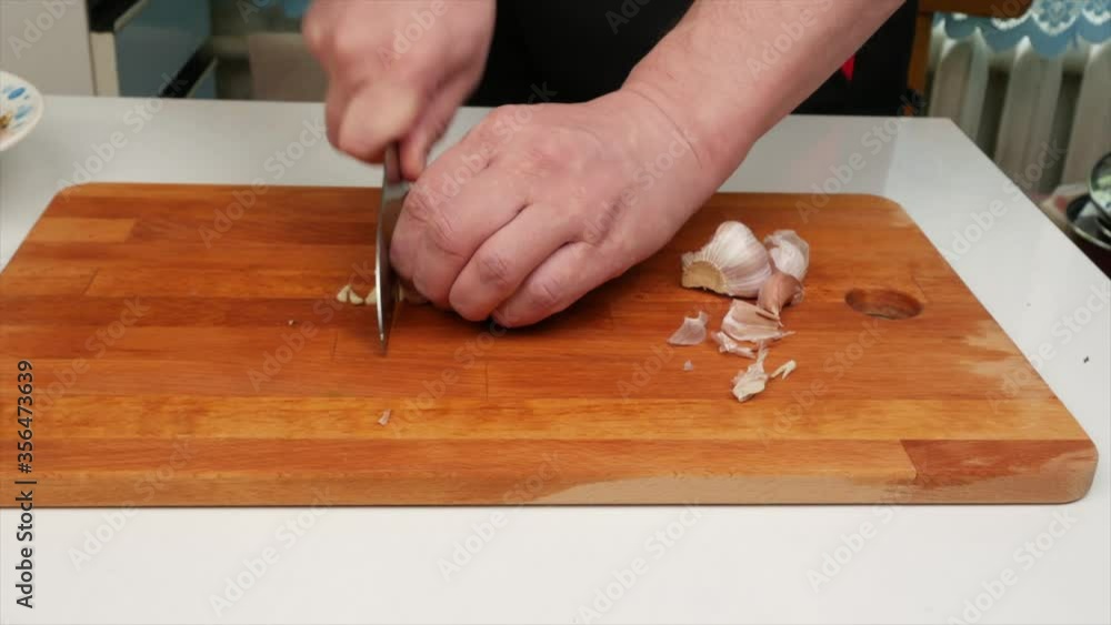 A man peels and cuts garlic with a knife on a wooden kitchen board, hands, close-up A man peels and cuts garlic with a knife on a wooden kitchen board, hands, close-up