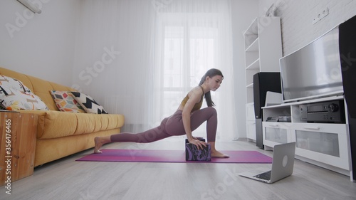 On-line work out woman using internet services with help of her instructor. Woman doing stretching exercises on violet mat and looking on the notebook