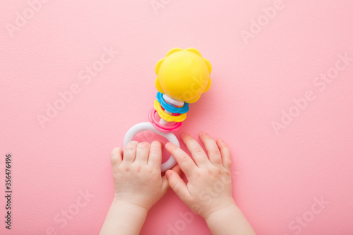 Baby girl hands playing with colorful rattle on light pink floor background. Pastel color. Closeup. Toys of development for infant. Point of view shot. Top down view. photo