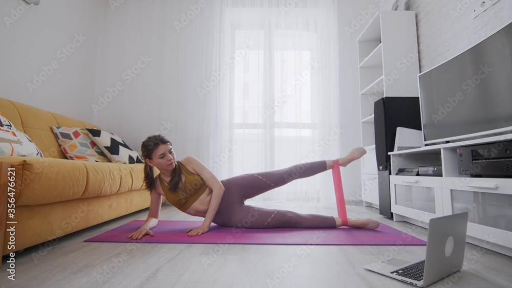On-line work out woman using internet services with help of her instructor. Woman lays on mat and lifts leg with resistance band On-line work out woman using internet services with help of her instructor. Woman lays on mat and lifts leg with resistance band