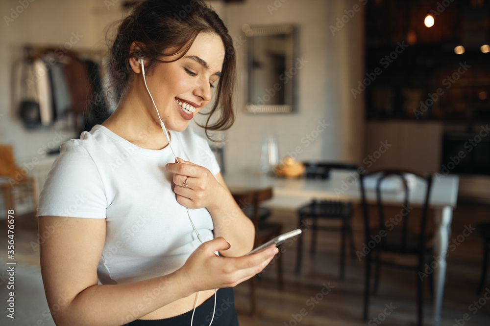 Indoor shot of charming young plus size woman in white top holding smart phone using headset while talking to friend via online video chat, smiling, making plans for weekend. People and technology - obrazy, fototapety, plakaty 