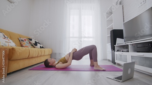 On-line work out woman using internet services with help of her instructor. Slim woman stands in bridge pose and lifts up dumbbell