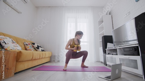 On-line work out woman using internet services with help of her instructor. Slim woman does classic squats with dumbbells