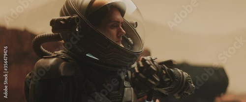 Caucasian female astronaunt checking hud on her suit while exploring planet surface, Mars rover in the background
