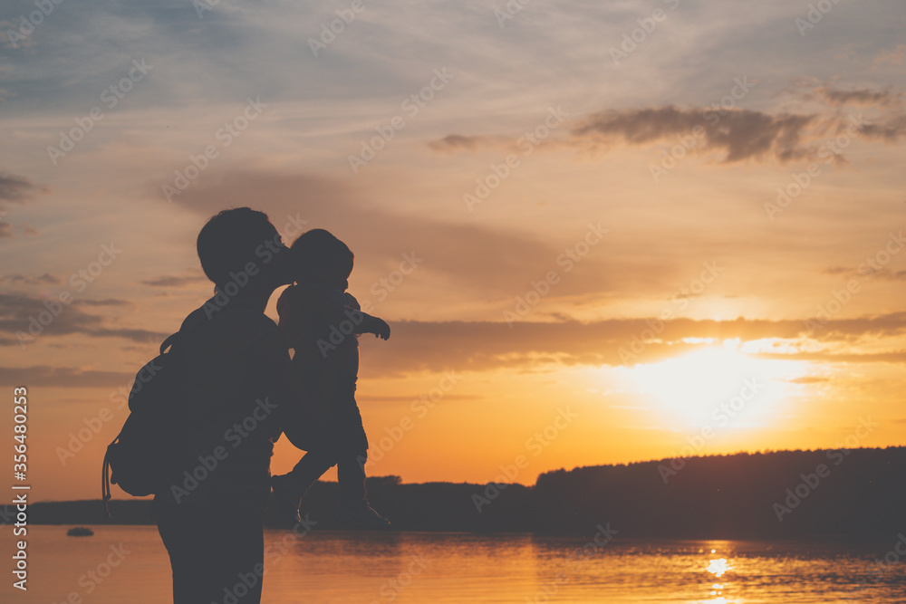 A sillhouete of mother and her baby playing together on the river side during sunset. Mom holding her toddler in hands and kissing its head.