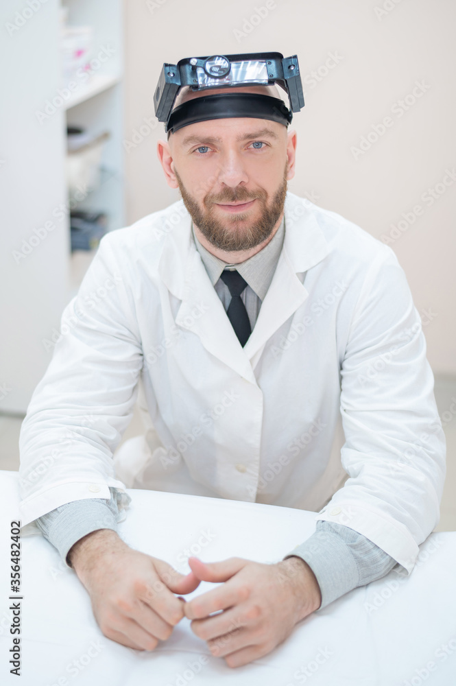 Male bearded doctor in a medical coat and with a magnifying head strap w lights sitting on the desk. Optometrist equipment. Medical office. Male bearded doctor in a medical coat and with a magnifying head strap w lights sitting on the desk. Optometrist equipment. Medical office.