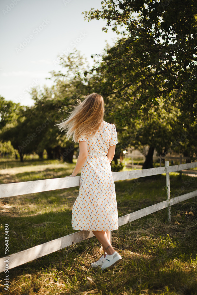 Naklejka premium Girl in a white dress walking in nature. Blonde in motion on a farm. A young girl enjoy the summer. 