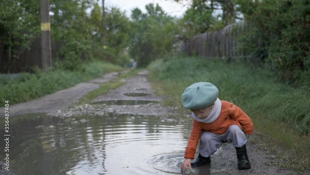 happy carefree male baby in a hat and rubber boots have fun playing with leaves in puddle on road after rain background of green bushes happy carefree male baby in a hat and rubber boots have fun playing with leaves in puddle on road after rain background of green bushes
