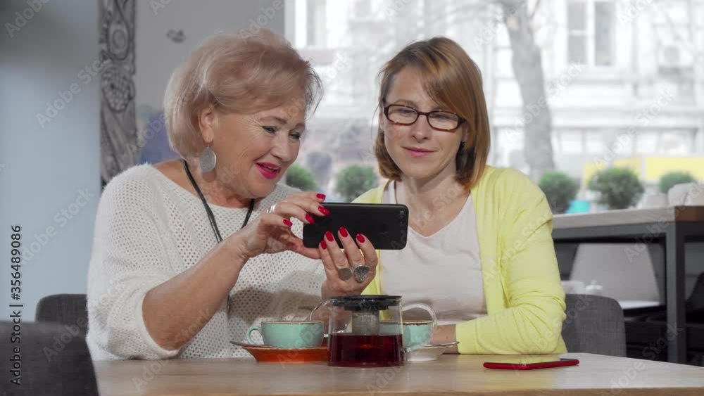 Mature woman and her senior mother using smart phone at the cafe together. Elderly woman showing something online on her phone to her adult daughter. Mobile app concept Mature woman and her senior mother using smart phone at the cafe together. Elderly woman showing something online on her phone to her adult daughter. Mobile app concept